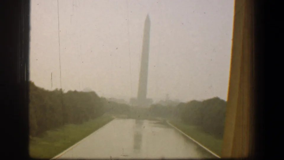 1961 Washington, DC home-movie frame showing the Washington Monument beyond the Reflecting Pool, with trees and window framing visible.