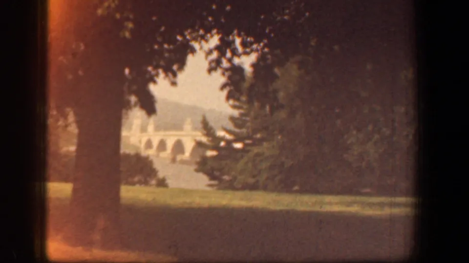 Warm-toned 1961 Washington, DC frame showing a shaded tree line, open grass, and a distant multi-arched bridge through the foliage.