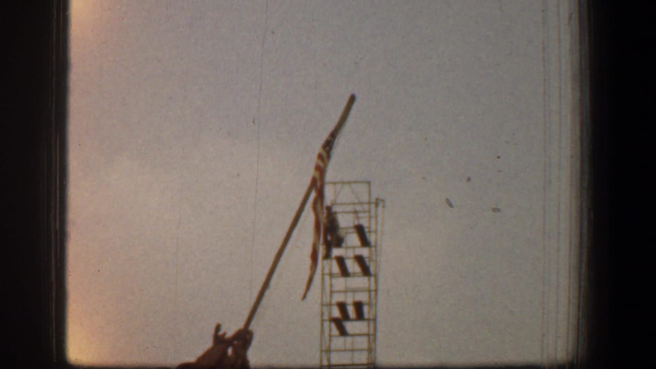 Low-angle view of the flagpole, flag, scaffolding, and partial soldier monument against a pale sky.