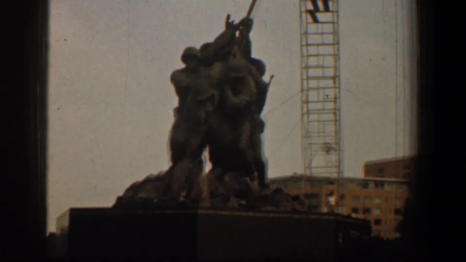 A closer 1961 home-movie view frames the memorial figures and base, with a crane tower and brick building in the Washington DC background.