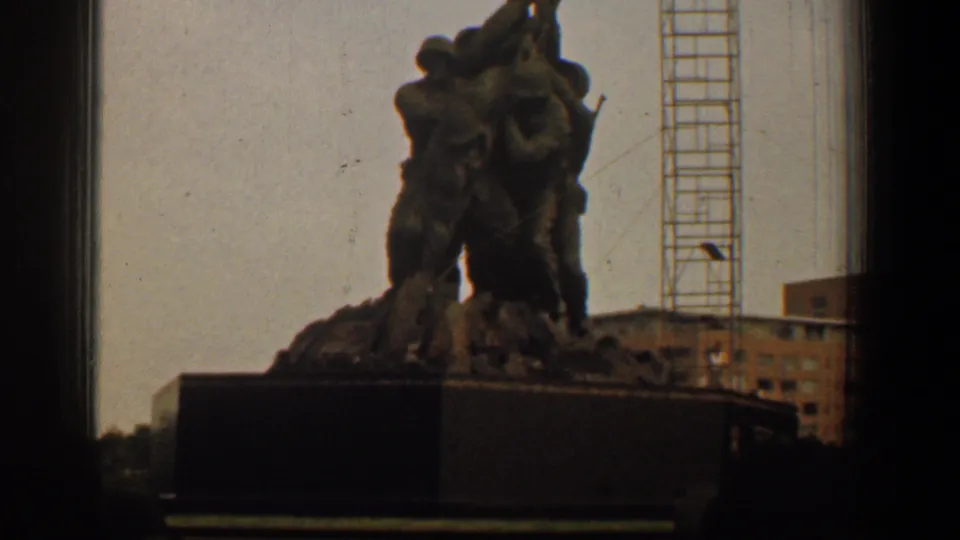 The final frame shows the Iwo Jima Memorial on its dark base, with cloudy sky, a crane, and a brick building behind the landmark.