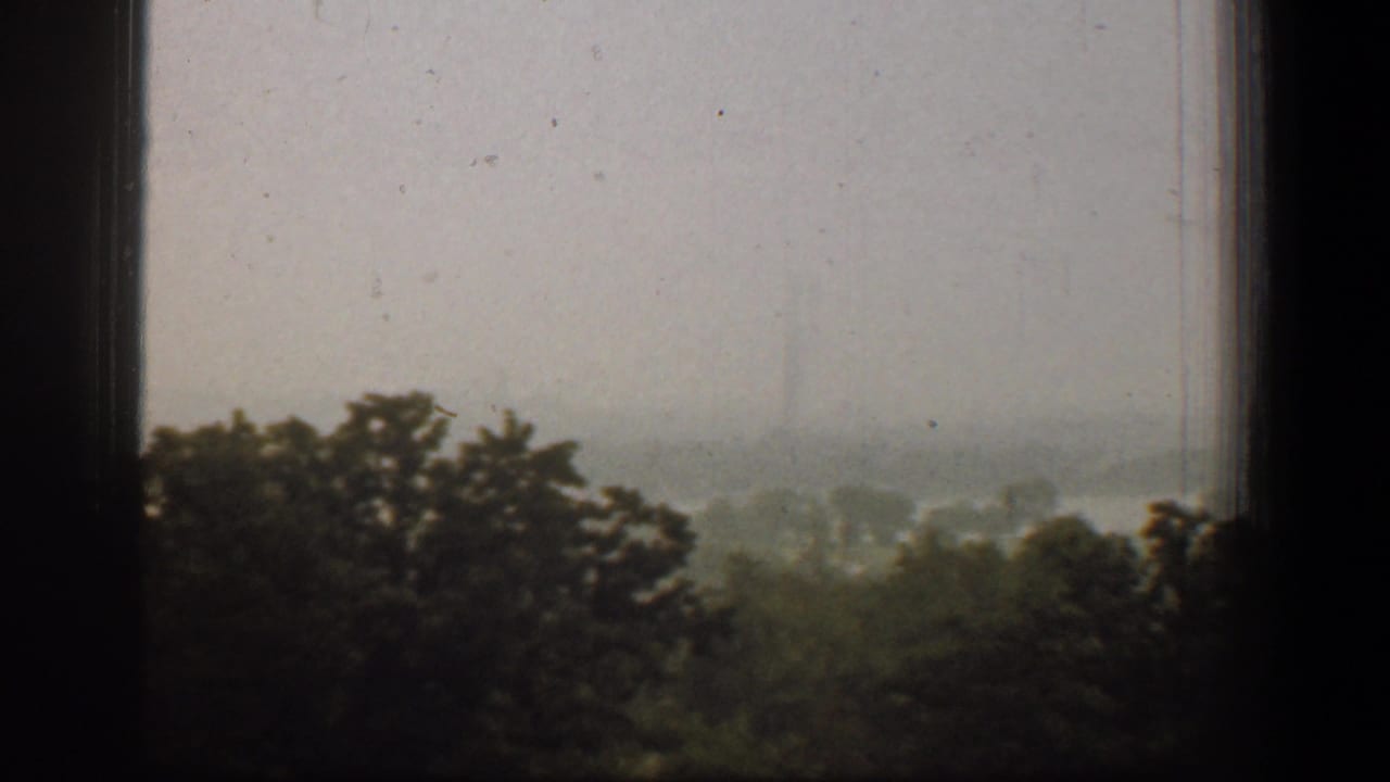 1961 Washington, DC home-movie frame showing mist-covered trees through a dark window frame, with a pale grey sky beyond.
