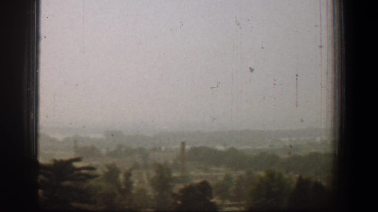 1961 Washington, DC home-movie view through dark window panes toward a hazy treeline and distant buildings.