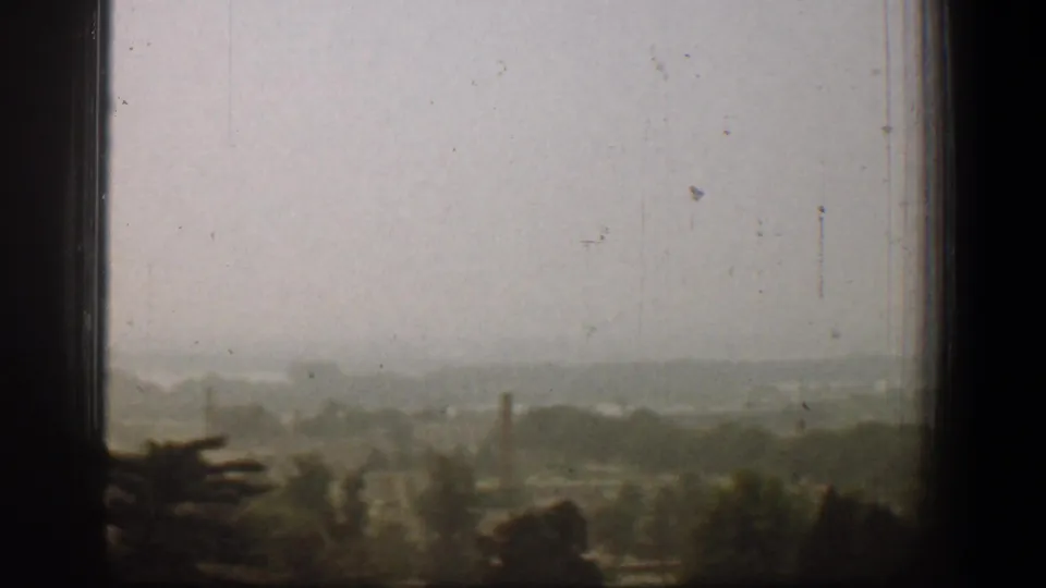 1961 Washington, DC home-movie view through dark window panes toward a hazy treeline and distant buildings.