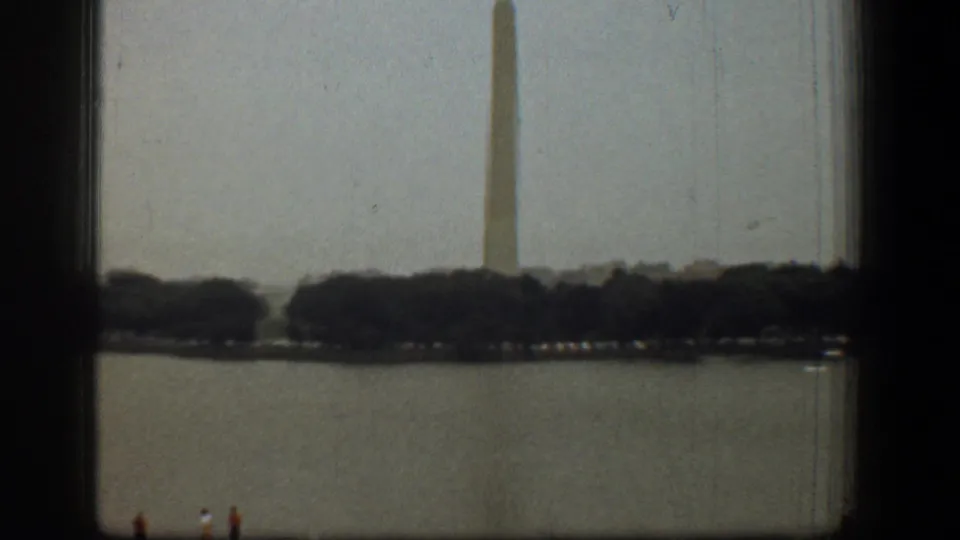 Soft 8mm frame of the Washington Monument rising behind trees, with the Reflecting Pool in front and small visitors at the lower edge.