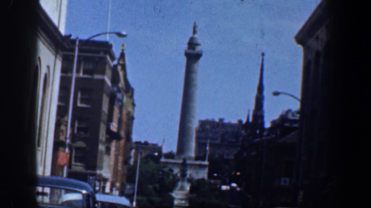 1961 home-movie street view in Washington, DC, showing parked cars, buildings, blue sky, and a distant monument column beyond the street.