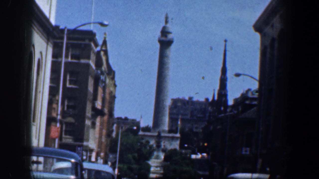 Downtown street view with a tall column monument, brick buildings, cars, and a church steeple.