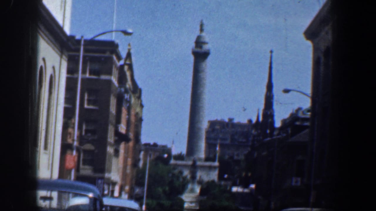 Washington, DC downtown view with a column monument, church steeple, streetlamps, brick buildings, and early 1960s automobiles.
