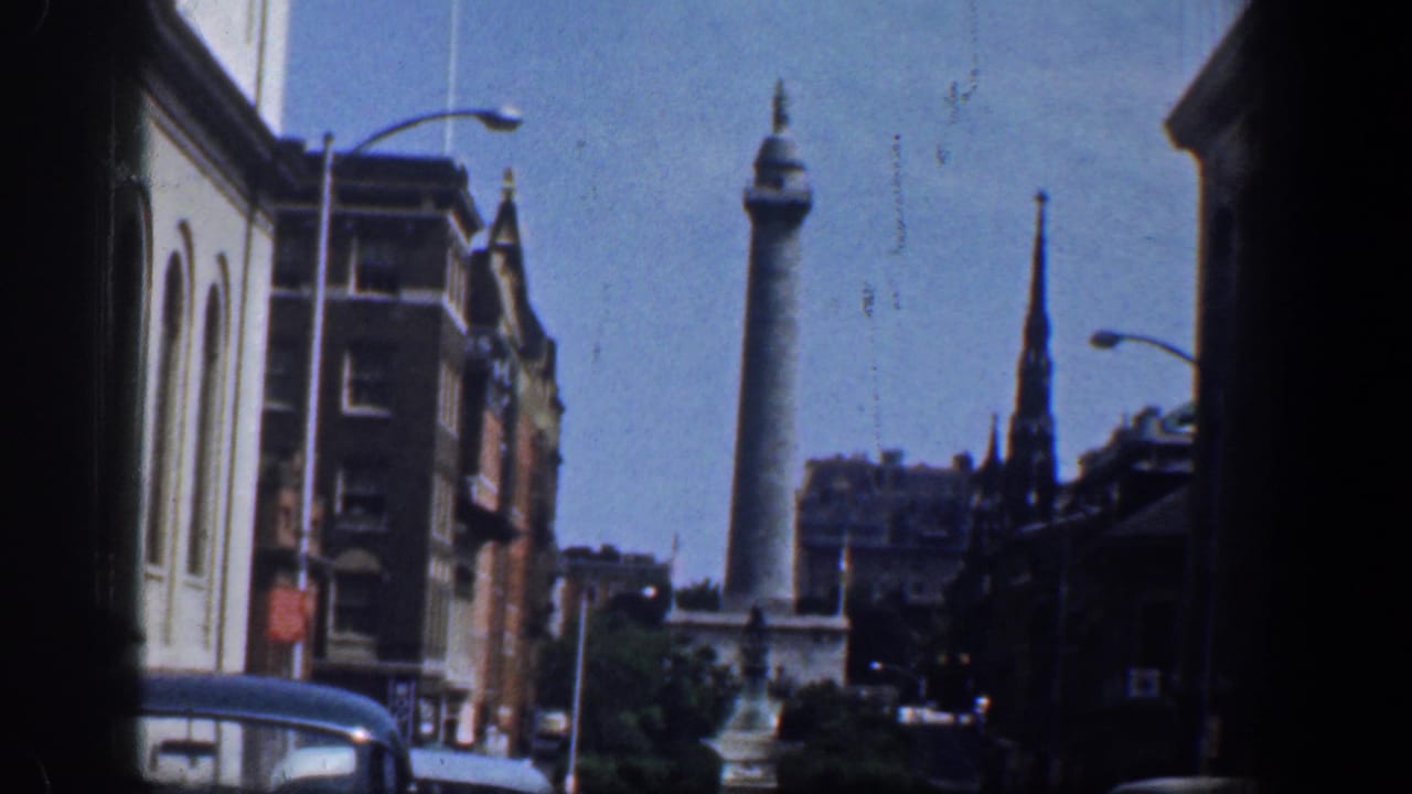 Soft 8mm frame of a Washington, DC downtown street, with a civic column monument, steeple, cars, buildings, and shadowed window edges.