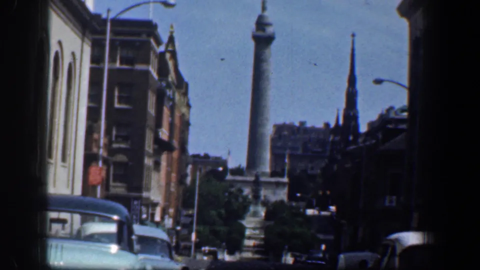 1961 Washington, DC downtown traffic scene with a tall monument, church steeple, masonry buildings, streetlamps, and period automobiles.