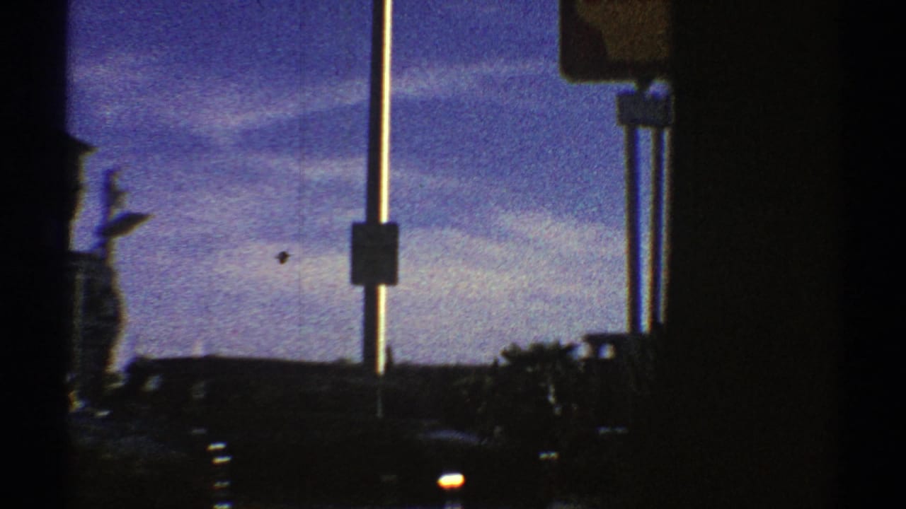 Moving 1969 Nevada street view partly blocked by vehicle window frames, with roadside signs, cars, poles, and distant buildings.