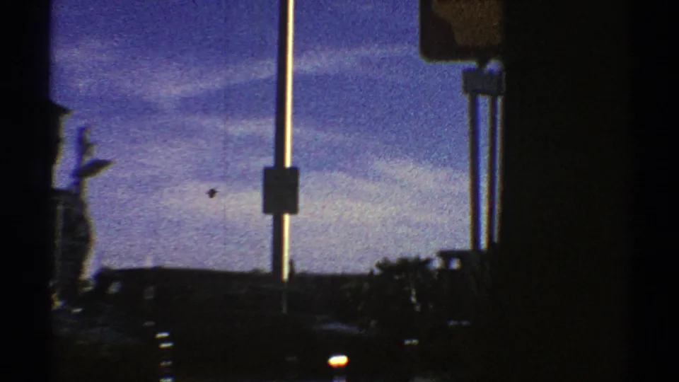 Moving 1969 Nevada street view partly blocked by vehicle window frames, with roadside signs, cars, poles, and distant buildings.
