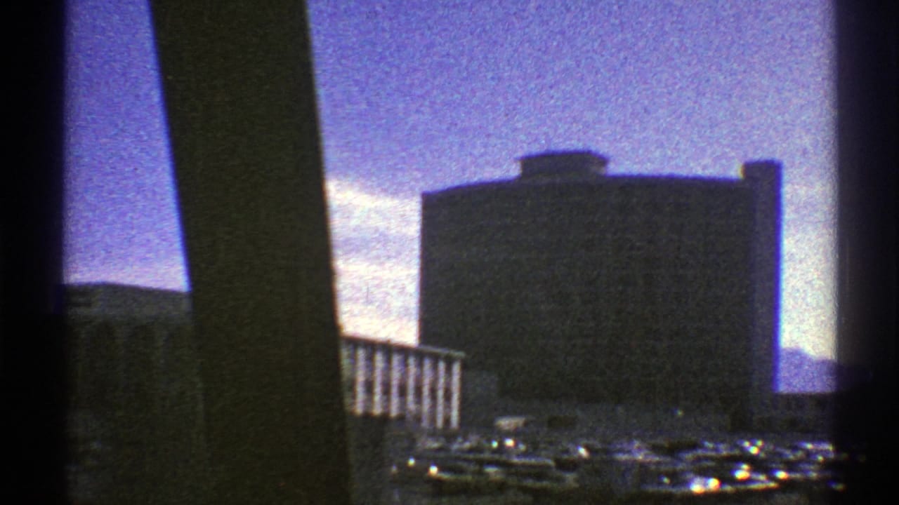 End of the 1969 Nevada moving city view, showing a large rectangular building, parking area, cars, and dark window framing.