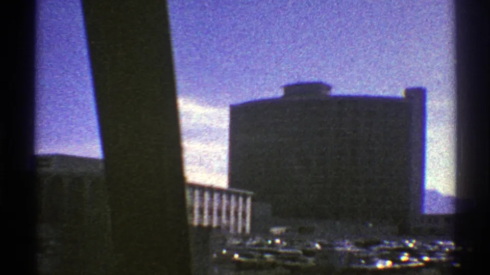End of the 1969 Nevada moving city view, showing a large rectangular building, parking area, cars, and dark window framing.