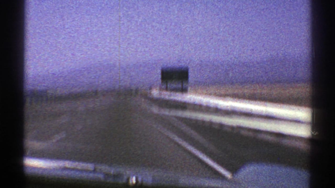 Moving-car view on a Nevada highway with guardrail, open road, and a distant sign under hazy 8mm color shift.