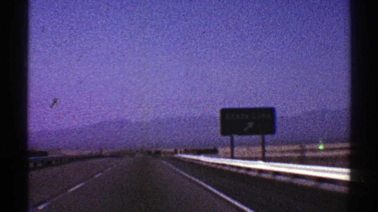 1969 Nevada road-trip frame showing a highway receding toward a sign and low mountains in grainy 8mm color.