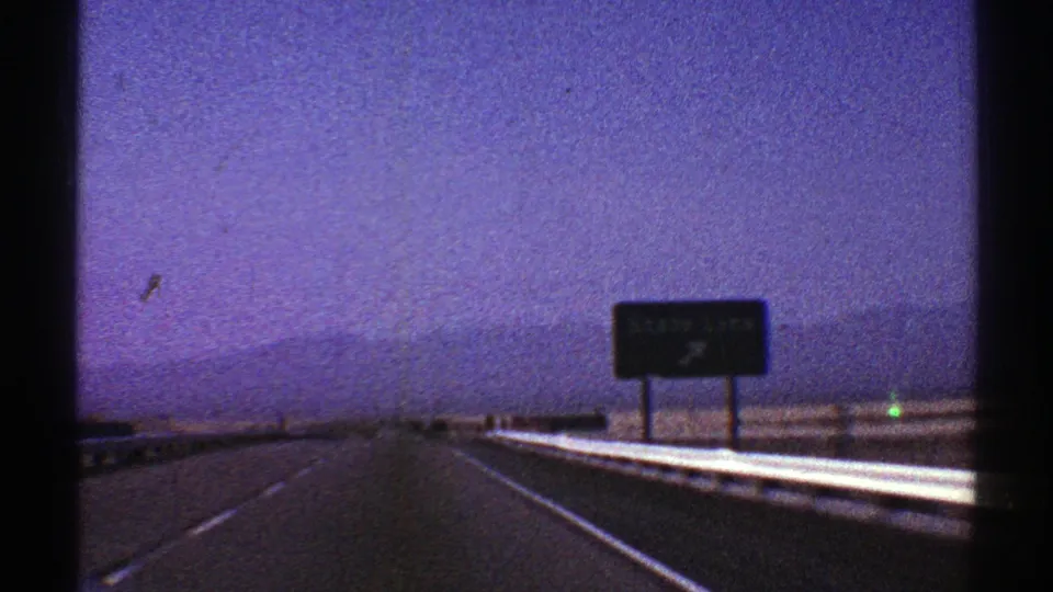 1969 Nevada road-trip frame showing a highway receding toward a sign and low mountains in grainy 8mm color.