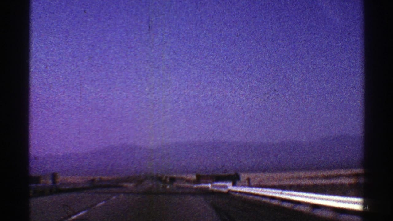 Desert highway seen from a moving vehicle with distant mountains and a purple-blue sky.