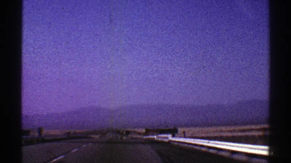 1969 Nevada 8mm view from a moving vehicle on a divided desert highway, with guardrails, distant mountains, and a hazy purple-blue sky.