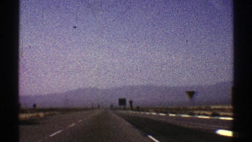 1969 Nevada road footage showing a straight desert highway, right-side road signs, pale guardrail highlights, and mountains under a hazy sky.