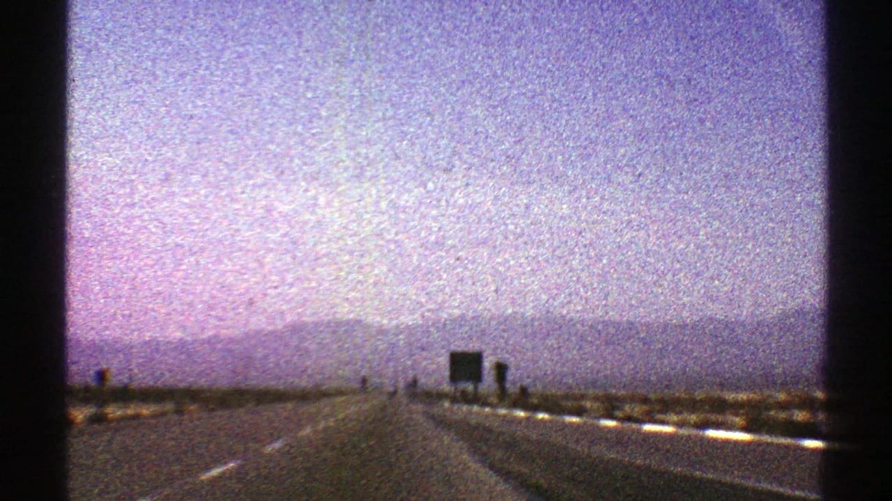 1969 Nevada 8mm frame of an open highway approaching a dark roadside sign, with desert shoulders and distant mountains below a washed lavender sky.