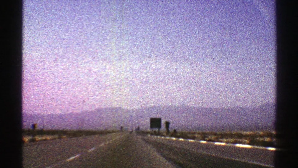 1969 Nevada 8mm frame of an open highway approaching a dark roadside sign, with desert shoulders and distant mountains below a washed lavender sky.