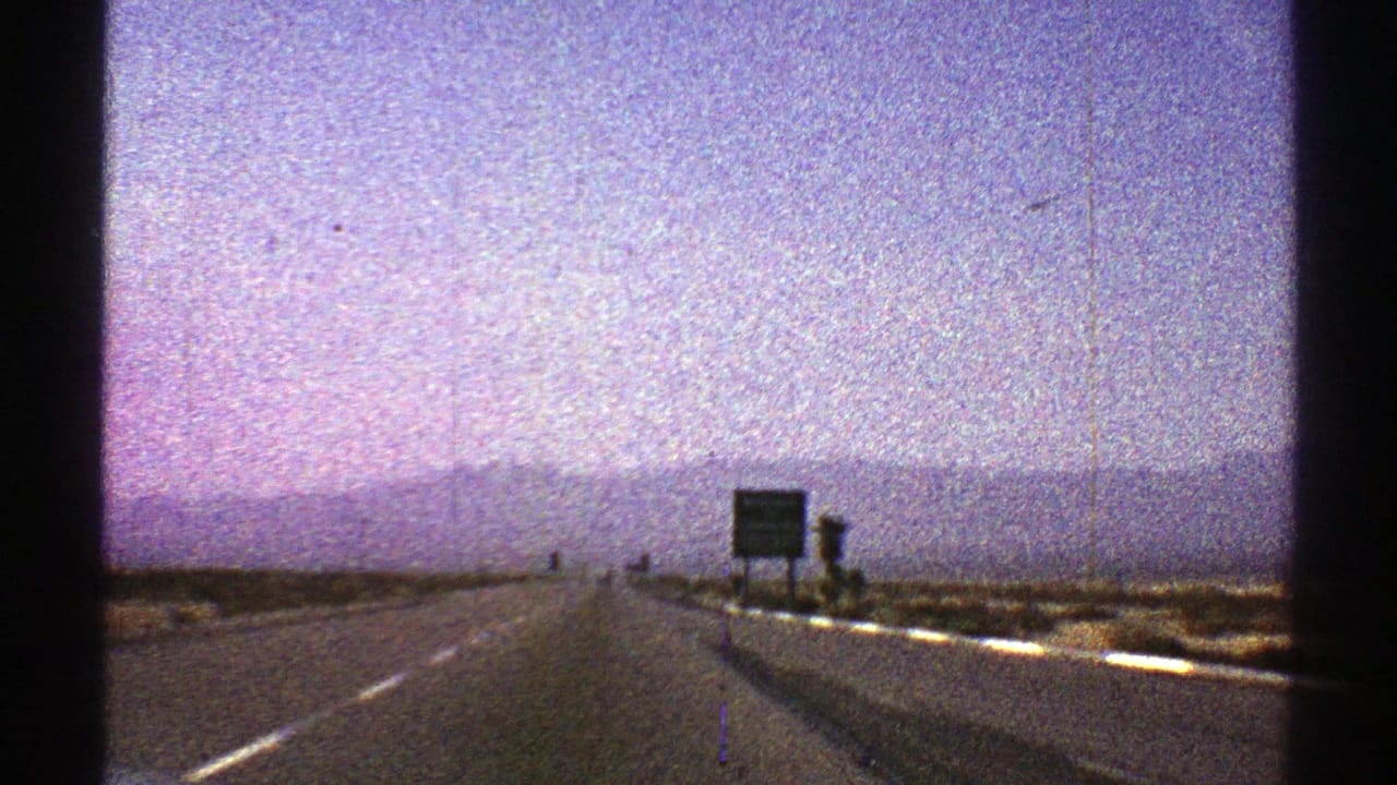 1969 Nevada highway view with roadside signs near the right shoulder, a straight lane ahead, distant mountains, and visible 8mm film grain.
