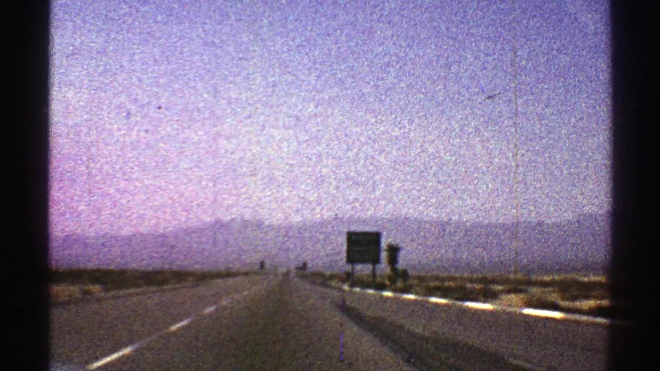 1969 Nevada highway view with roadside signs near the right shoulder, a straight lane ahead, distant mountains, and visible 8mm film grain.