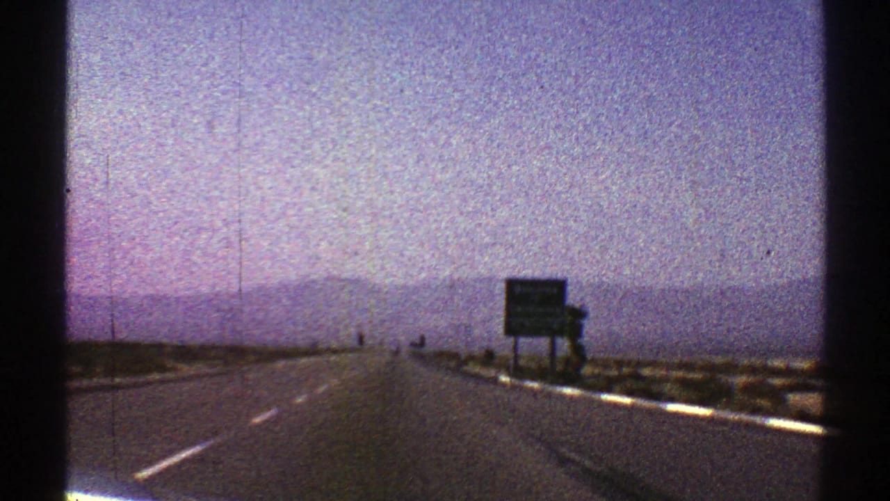 Straight desert highway with distant mountains and roadside signs under a hazy sky.