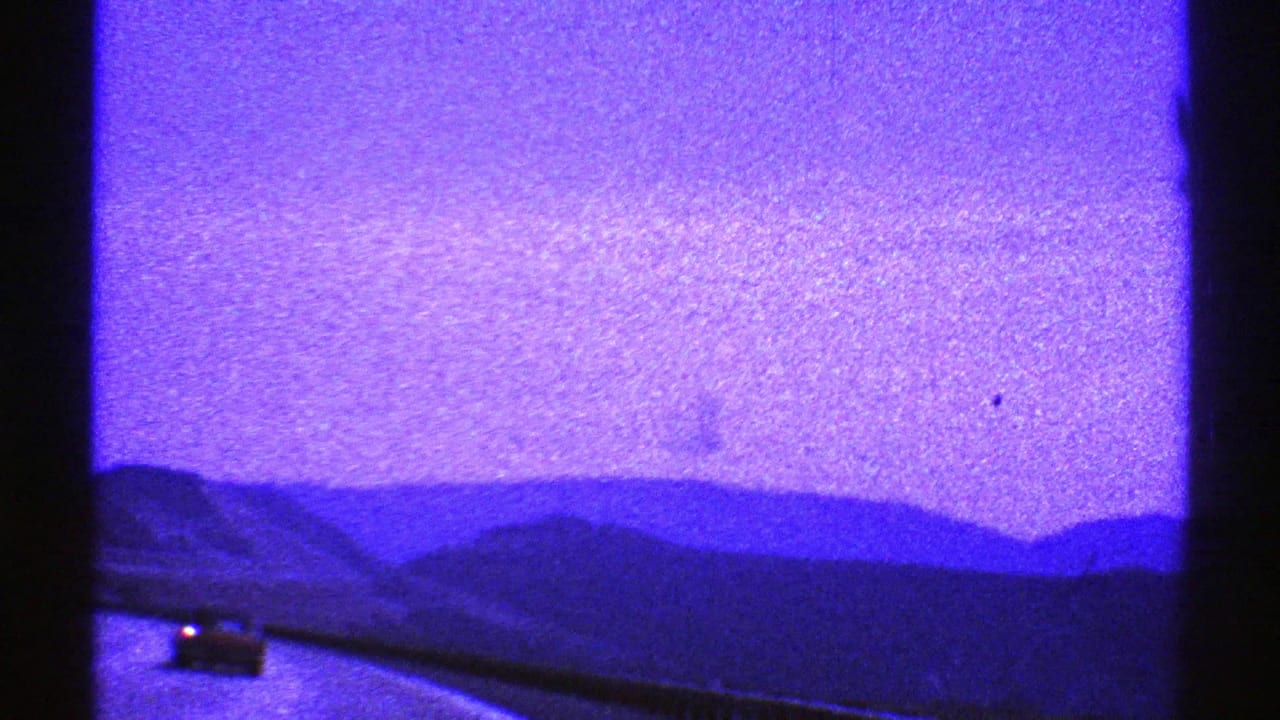 A Nevada highway recedes toward low mountains, seen through a dark window divider with one distant car on the road.