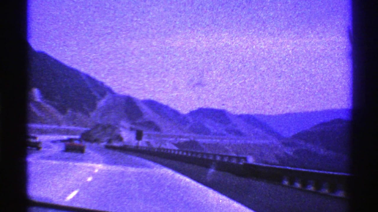 1969 Nevada road-trip frame showing cars ahead on a curving mountain highway, roadside guardrail, hills, and purple 8mm color shift.