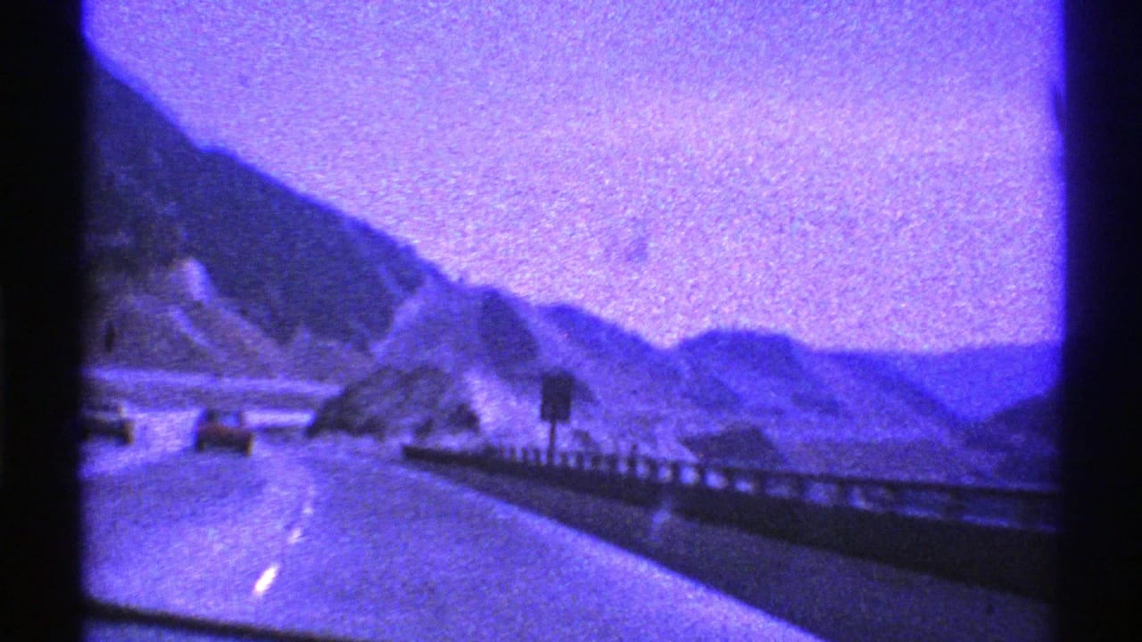 1969 Nevada 8mm frame of a moving highway view with cars ahead, a roadside sign, guardrail, rocky hills, and a broad purple sky.