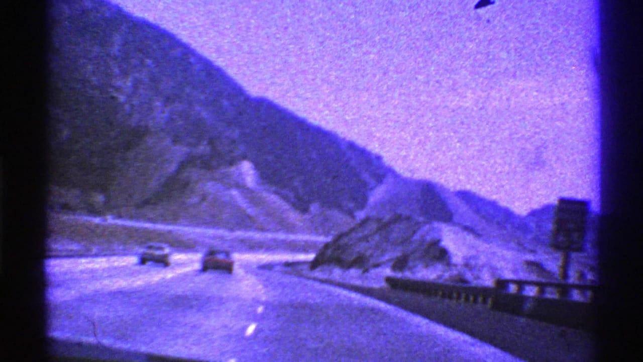 1969 Nevada 8mm road scene with a curving paved highway, two cars ahead, rocky mountains, guardrail, sign, and heavy purple color shift.