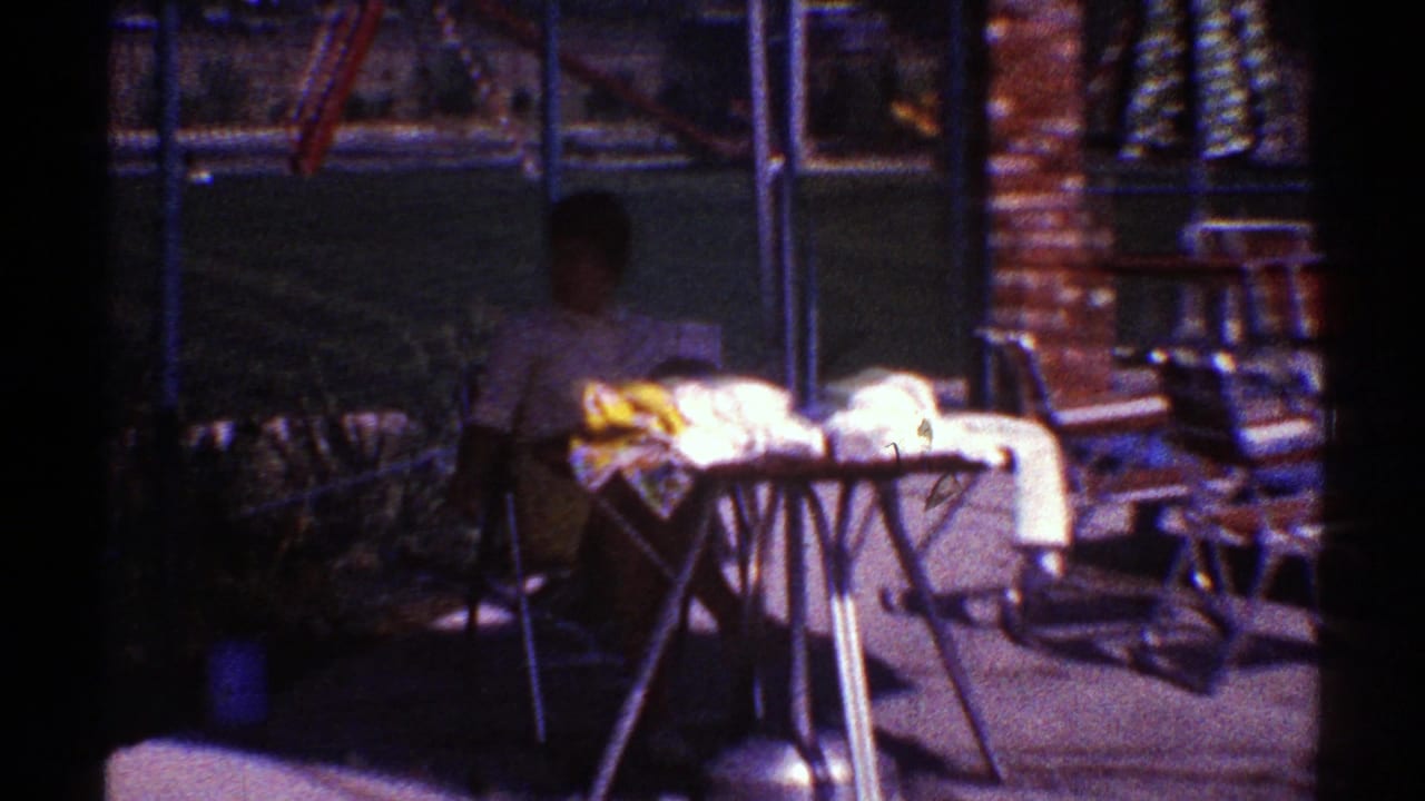 Woman seated outside near a covered table beside a brick wall and chain-link fence.