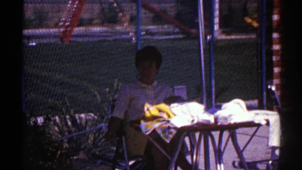 1969 Nevada frame of a woman seated outdoors near a cloth-covered table, with chain-link fence, brick storefront detail, plants, and a red ladder visible.
