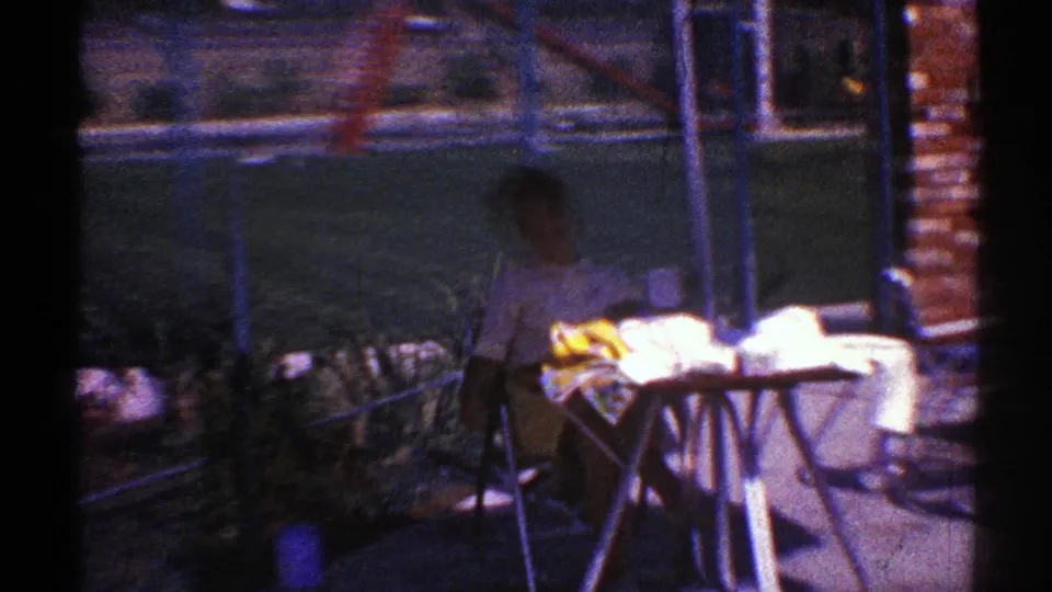 A shadowed 1969 Nevada home-movie view shows the seated woman beside the covered table, with brickwork, fencing, plants, and patio objects around her.