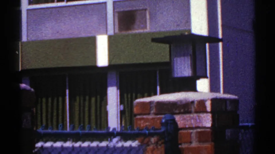 1969 Nevada 8mm frame of a green-and-white building exterior partly obscured by fencing, brickwork, and a lamp post.
