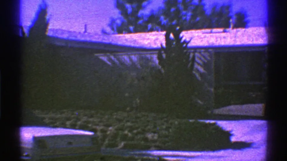 1969 Nevada home-movie frame with a low residential roofline, snow, shrubs, driveway, and trees in soft 8mm color.