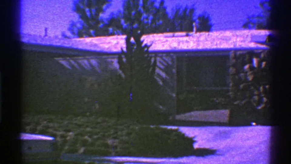 1969 Nevada 8mm frame showing a quiet suburban exterior with roof snow, dense shrubs, driveway, and stacked firewood.