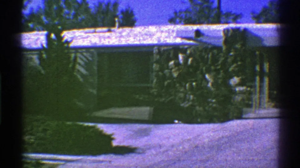 1969 Nevada home-movie frame of a driveway, walkway, shrubs, trees, and low suburban house with snow on the roof.