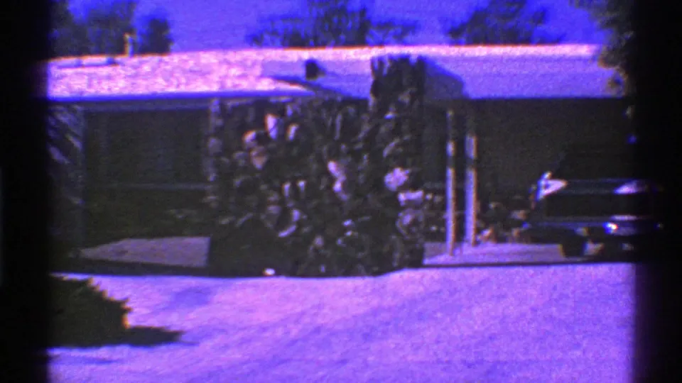 1969 Nevada home-movie frame showing a parked classic car in a suburban driveway near a low house, awning, shrubs, and trees.