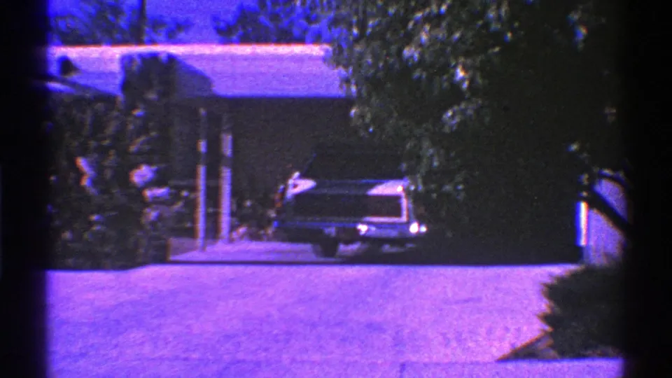 Final 8mm frame of the Nevada suburban exterior, with a classic car in the shaded driveway and trees partly obscuring the house.