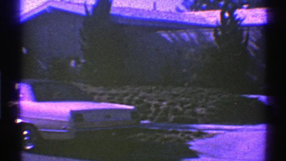 1969 Nevada frame showing the rear of a parked car near a single-story house, driveway, and dense residential landscaping.