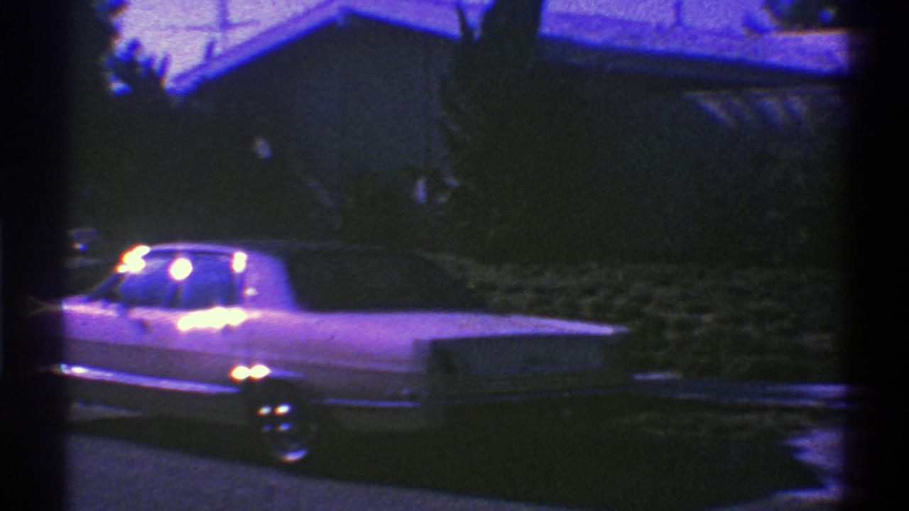 A 1969 Nevada 8mm frame shows a light-colored classic American sedan parked curbside at night, with bright lights reflecting against a dark residential backdrop.