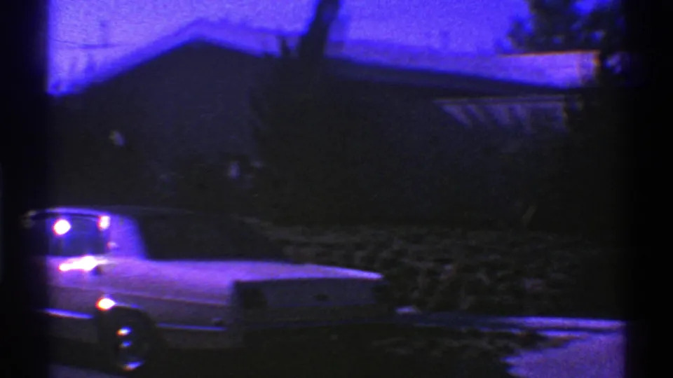 The 8mm frame catches the front portion of a light-colored classic car at the edge of the image, with dark shrubbery and a residential driveway nearby.