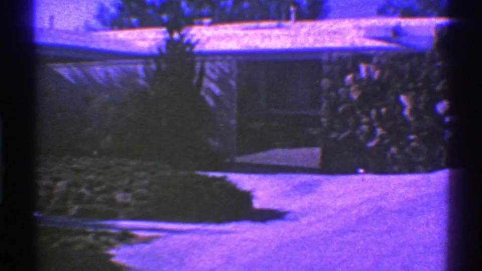 A nighttime 8mm frame shows a residential driveway, garage or house facade, landscaping, and purple-blue film cast, with the parked car no longer clearly visible.