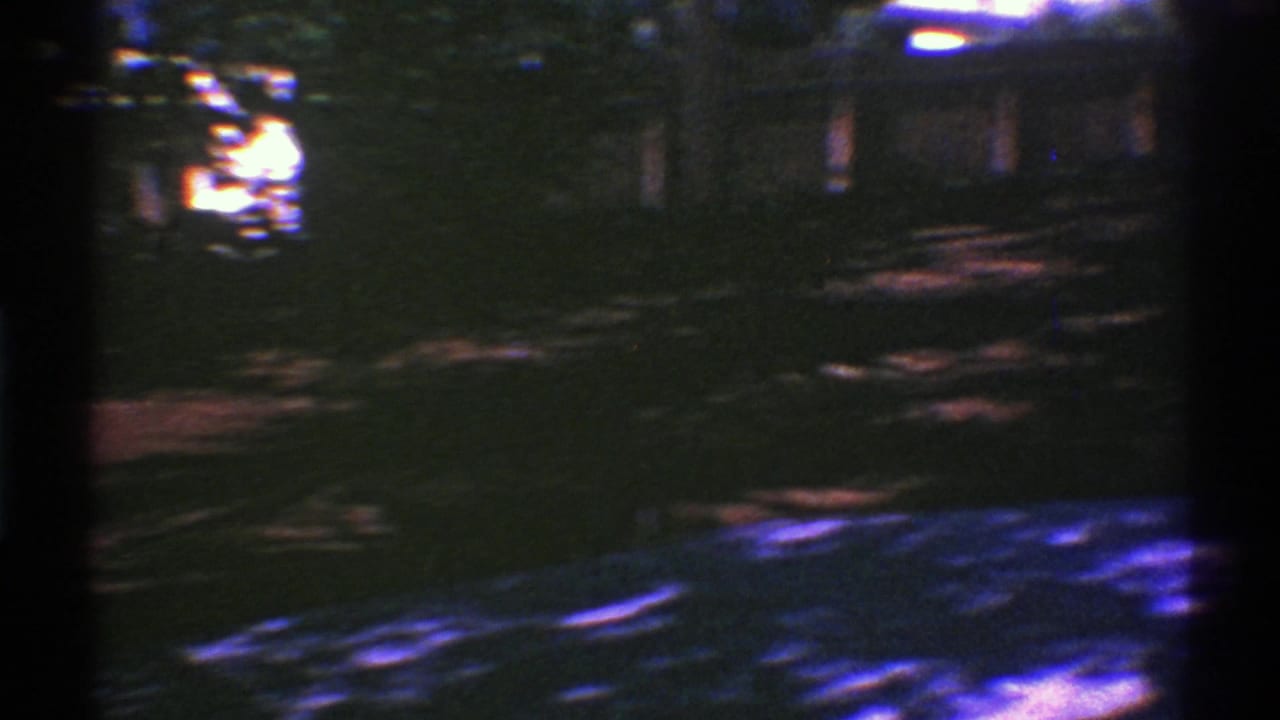 Grainy 1969 Nevada home-movie frame looking over a shaded yard toward trees and a dark fence or exterior structure.