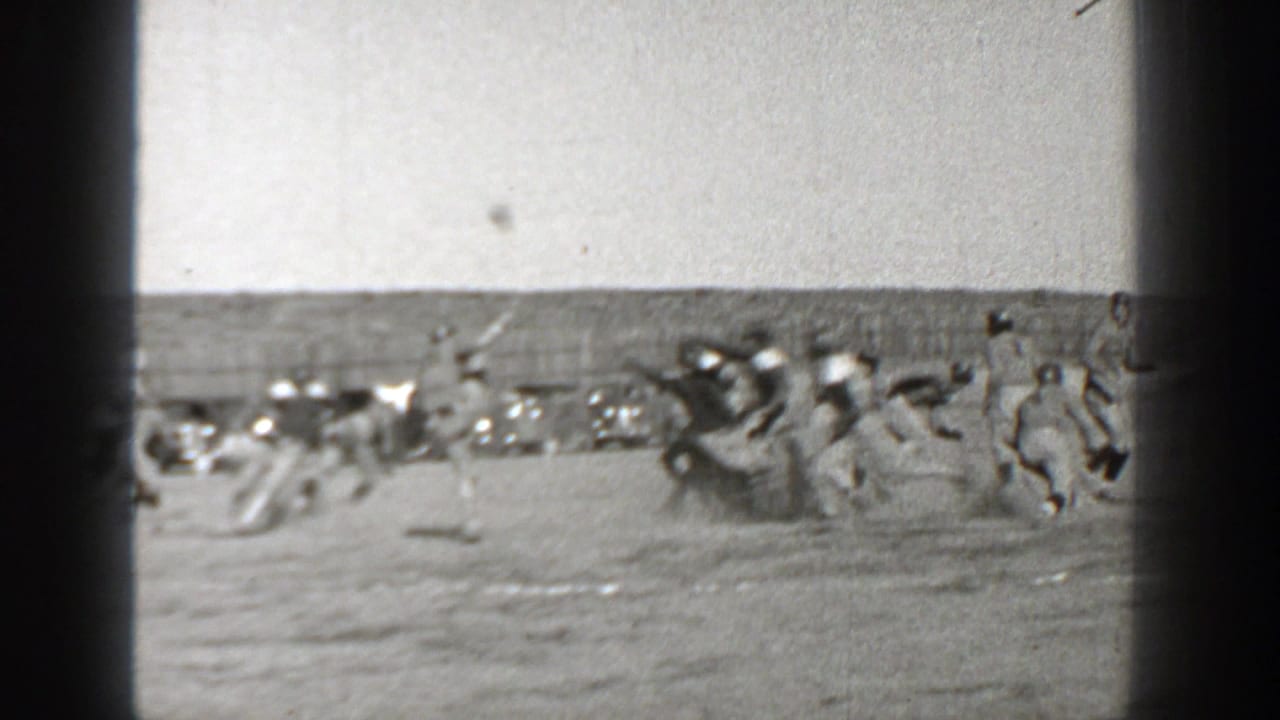 Football players cluster on a dusty field with parked cars behind the sideline.