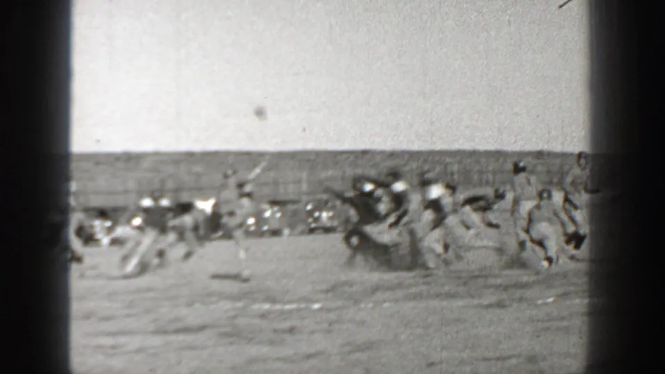 Washington 1938 home-movie frame showing a football play on a dusty field, with players in vintage uniforms and automobiles in the background.