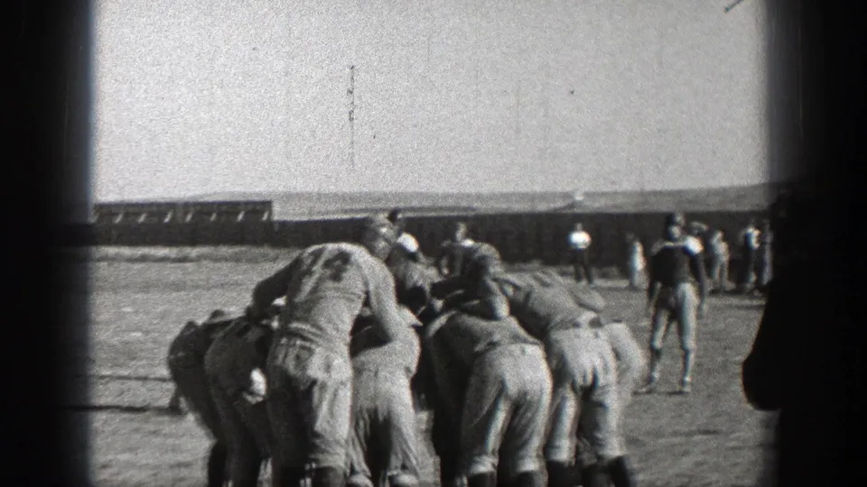 Washington 1938 football players in numbered uniforms and helmets cluster in a huddle on an outdoor field.